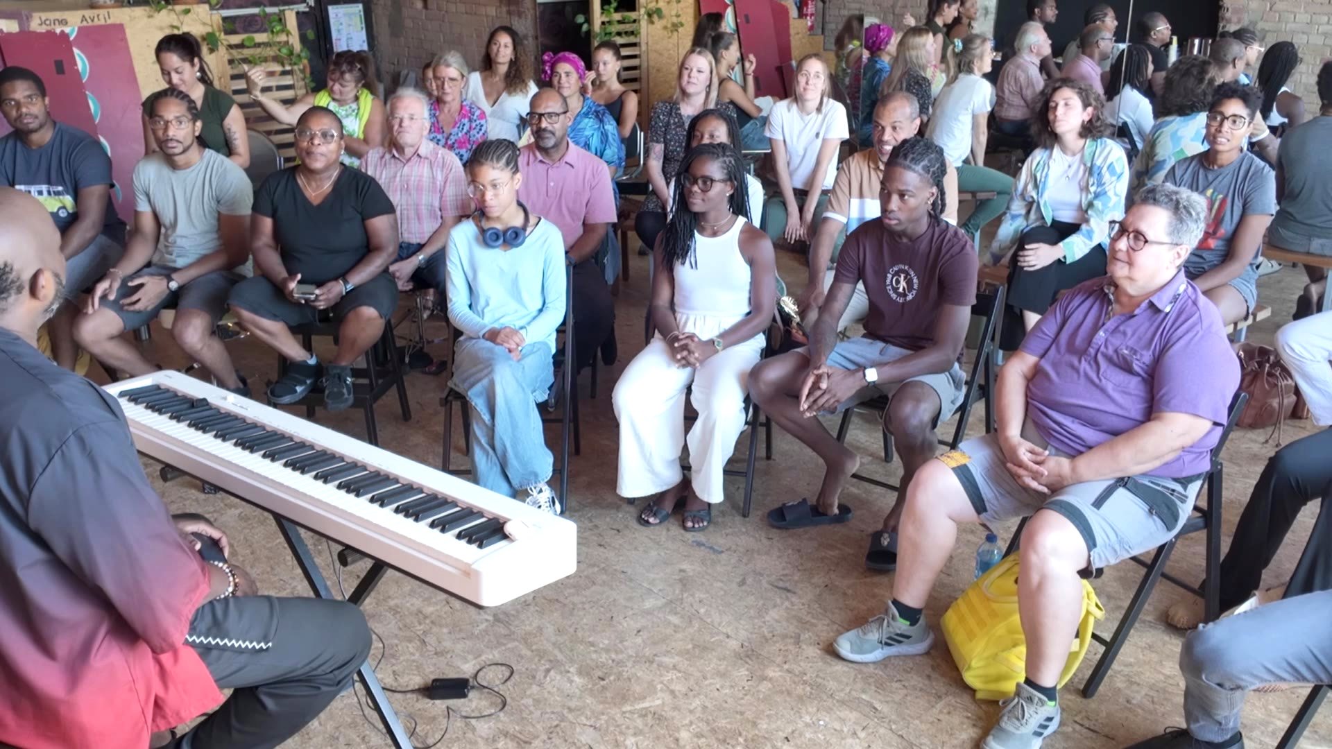 Caméra en plongée derrière Michel au piano, face aux participants de l'atelier Sum Voice